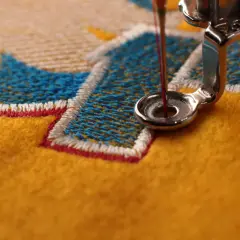 Gros plan sur une machine à coudre créant un point de broderie complexe sur un tissu jaune vif, avec des fils bleus et blancs formant un motif.