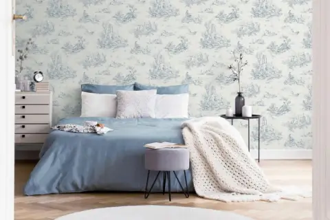 Chambre paisible avec un papier peint au motif jouy bleu et blanc, un lit bleu, des coussins blancs et une couverture en maille.