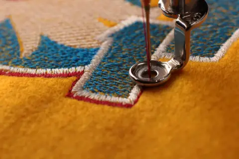 Gros plan sur une machine à coudre créant un point de broderie complexe sur un tissu jaune vif, avec des fils bleus et blancs formant un motif.