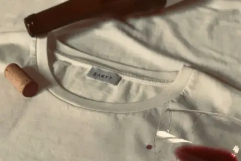 Un bouchon de liège et une bouteille de vin à côté d'un tee shirt blanc, avec une tache de vin rouge.