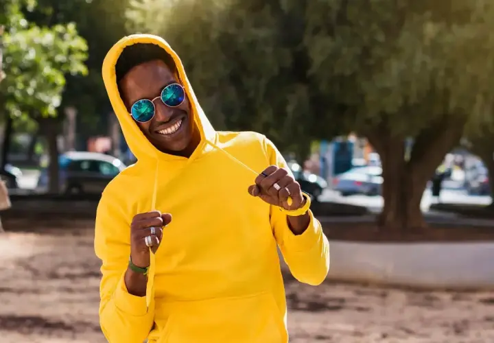 Un homme souriant porte un sweat jaune vif, ses lunettes rondes reflétant le soleil. Le grammage du sweat semble parfait pour une journée ensoleillée.