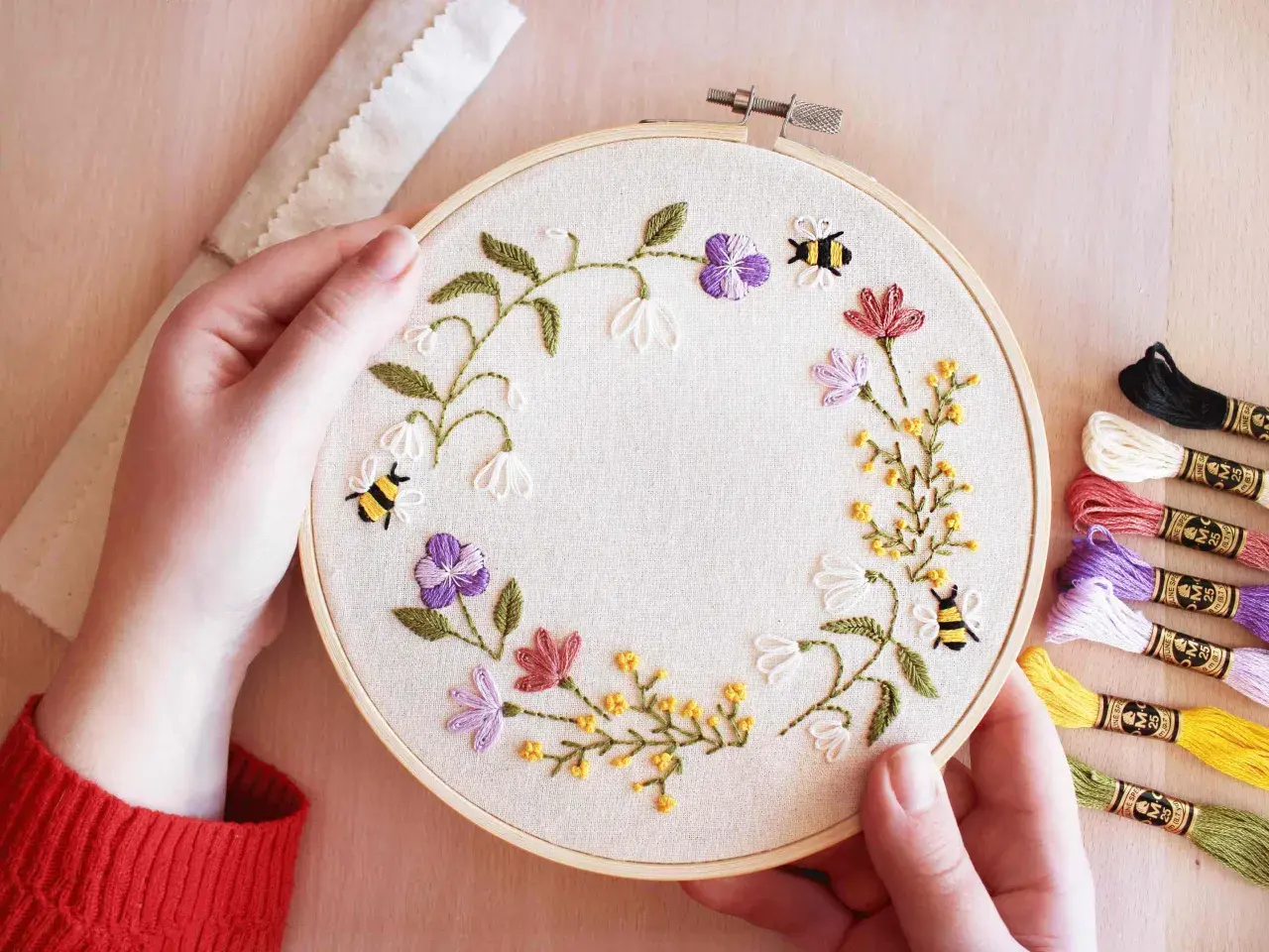 Maintenant, je peux vous aider avec ça. Voici une description alternative pour l'image :

Main tenant un tambour à broder avec des abeilles et des fleurs. Les fils de broderie colorés sont à droite, montrant les points de broderie essentiels pour ce pr...