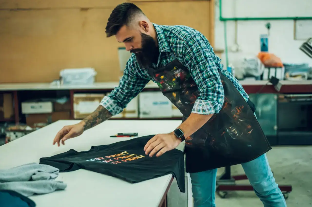 Un homme avec une barbe, portant un tablier tach&eacute; de peinture, v&eacute;rifie un t-shirt noir imprim&eacute; avec la technique DTG.