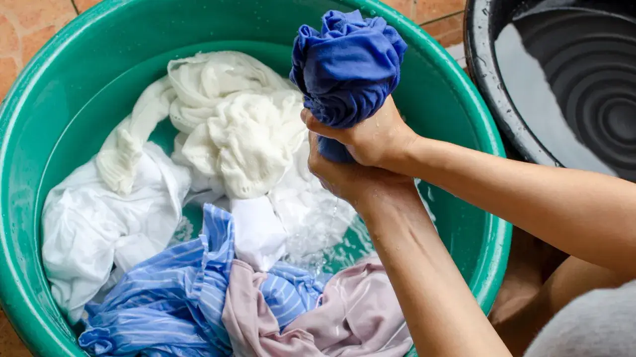 Mains tordant un vêtement bleu dans une bassine remplie de linge blanc et coloré. Un geste pour blanchir un vêtement.