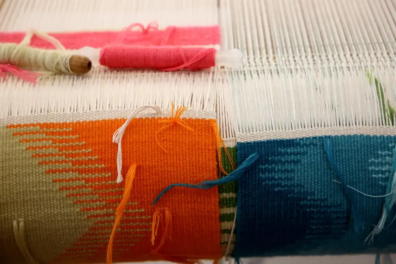 Sur un métier à tisser, des fils de laine aux couleurs vives (orange, bleu, rose) sont travaillés pour créer une toile d'Aubusson aux motifs géométriques.