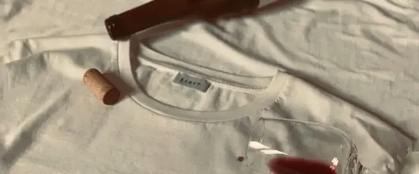 Un bouchon de liège et une bouteille de vin à côté d'un tee shirt blanc, avec une tache de vin rouge.